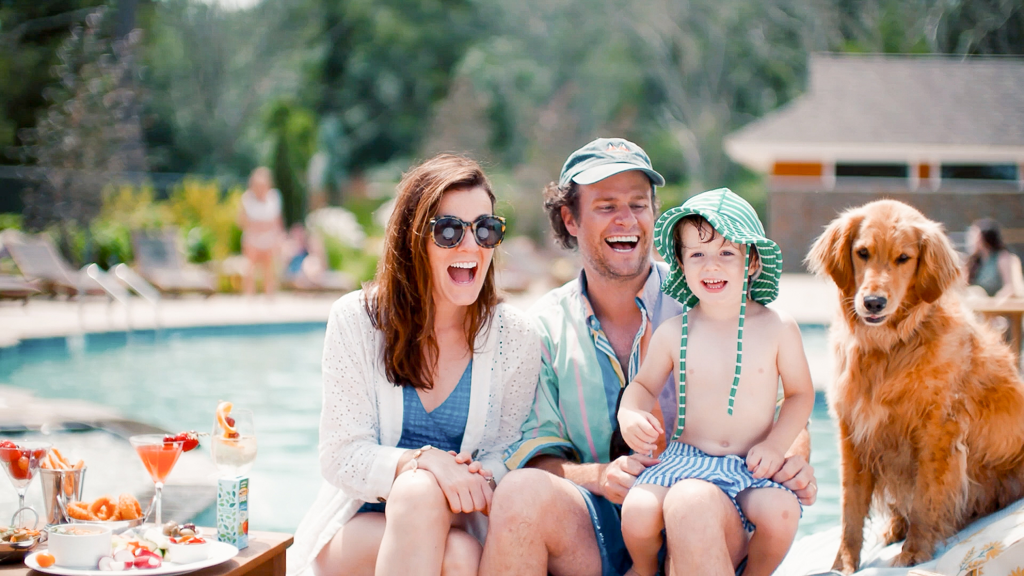A family of three, along with a golden retriever, is sitting by a pool with a table of drinks and snacks nearby, all smiling at the camera.