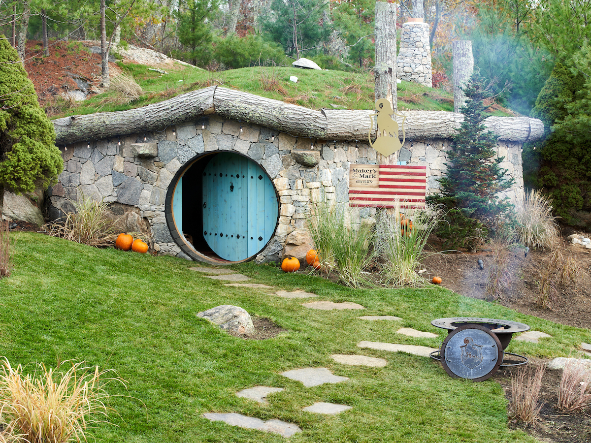 A stone house with a round, blue door and an American flag. The yard has pumpkins, stepping stones, and nearby trees.