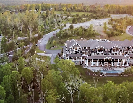 An aerial view of a large building with a pool, surrounded by dense trees and a winding road in a green landscape, is shown.