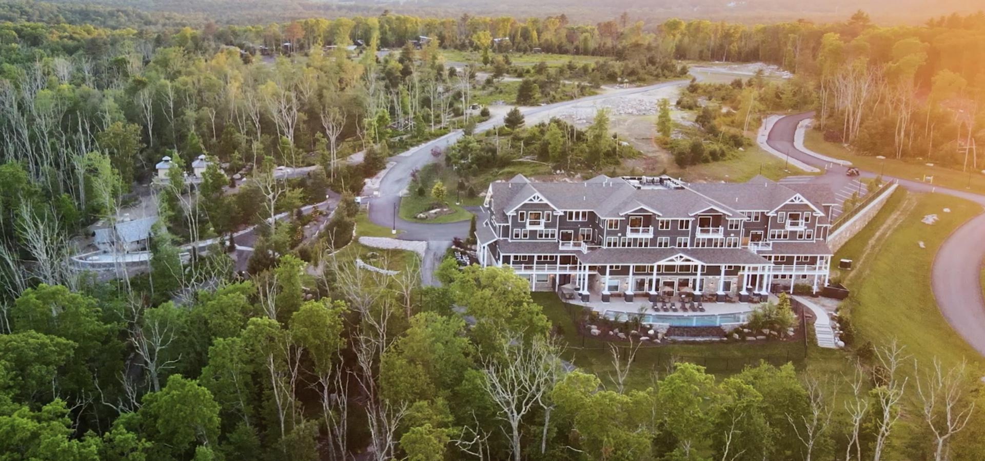 An aerial view of a large building with a pool, surrounded by dense trees and a winding road in a green landscape, is shown.
