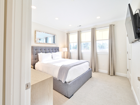 A modern bedroom with a bed, side table, lamp, and TV. Large windows with curtains provide natural light, creating a cozy atmosphere.