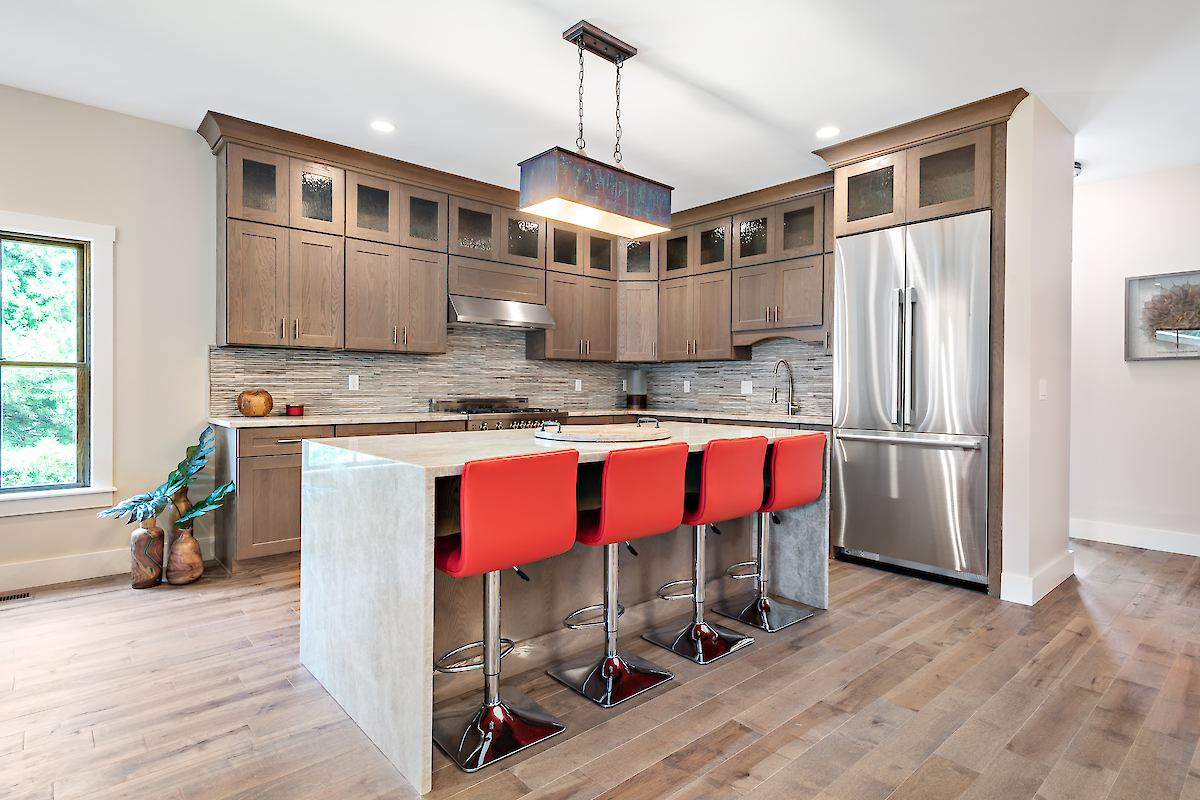 A modern kitchen with wooden cabinets, stainless steel appliances, a center island with red bar stools, and wooden flooring is shown.