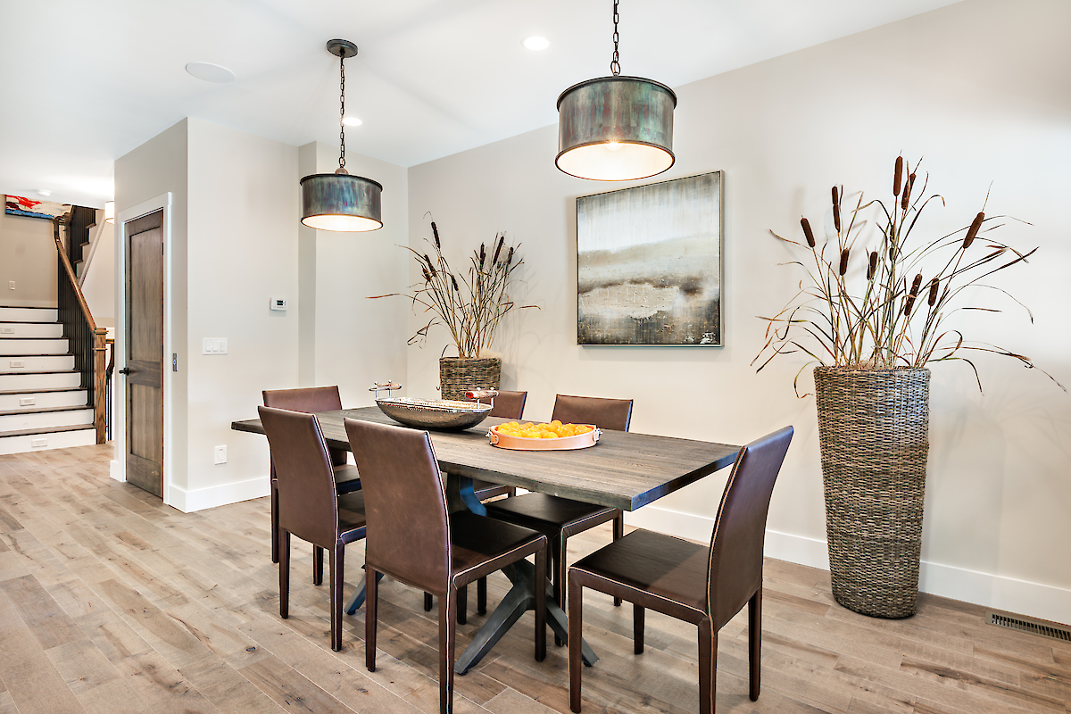 A modern dining room with a table set for six, two hanging lights, and large vases with decorative plants, ending the sentence.