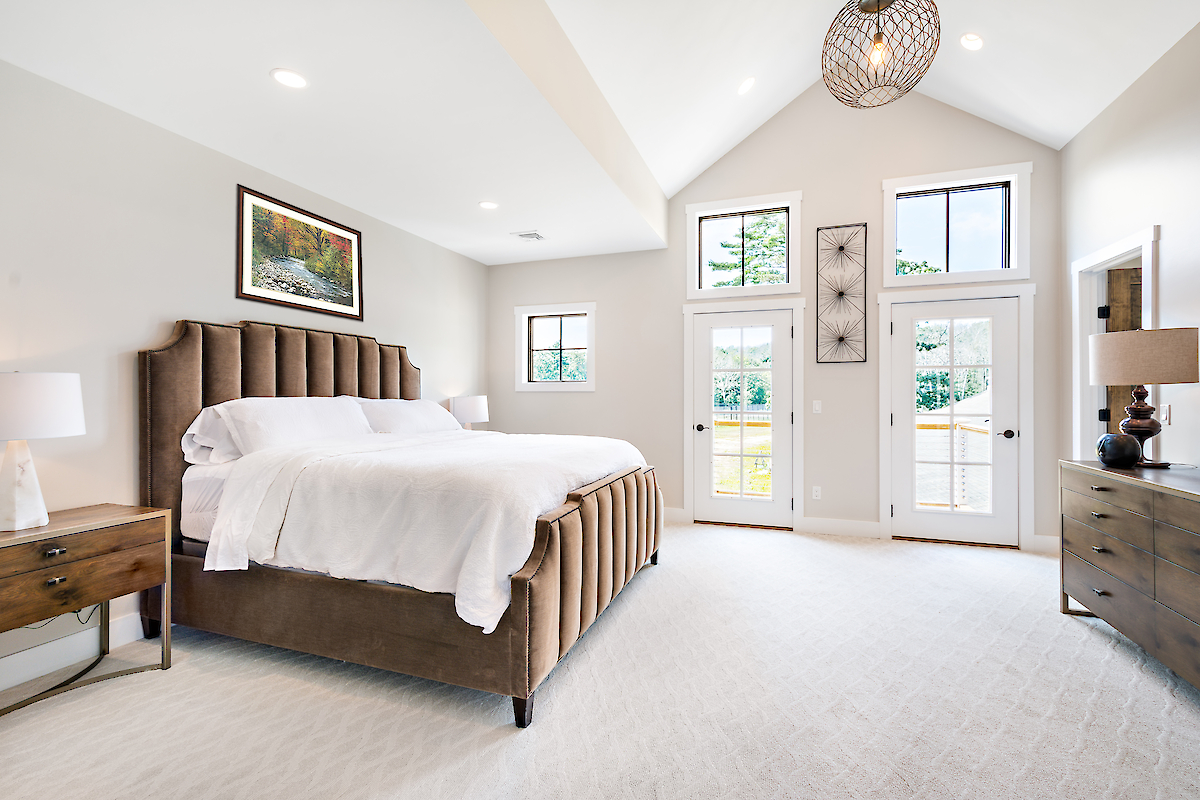 A spacious, well-lit bedroom with a large bed, wooden side tables, lamps, a painting, and large windows with doors leading outside.