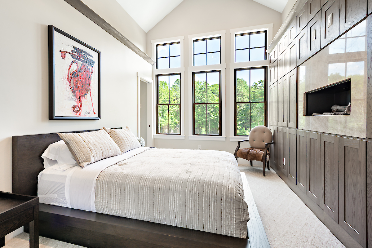 A modern bedroom features a large bed, four tall windows, a wall-mounted TV, an armchair, and artwork on the wall above the bed.