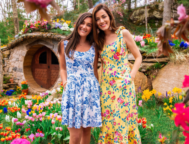 Two women in floral dresses posing in a colorful garden with flowers and a small round-door house in the background.
