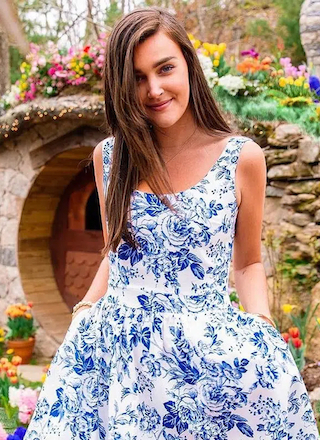 A woman in a floral dress stands in a colorful garden in front of a rustic stone house with a rounded door, surrounded by vibrant flowers.