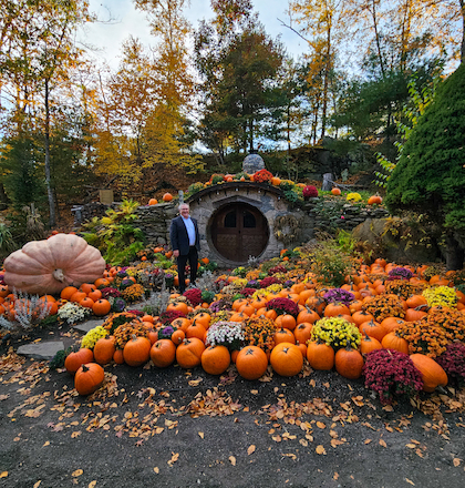Colorful pumpkins and autumn flowers surround a person standing near a round door set into a hillside, with trees in the background.