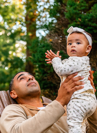 A man holds a baby outdoors, both looking upward with trees in the background. The baby is in patterned clothing with a headband.