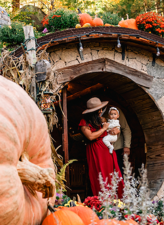 A person in a red dress and hat holds a baby at the entrance of a rustic, round door, surrounded by pumpkins and autumn decorations.
