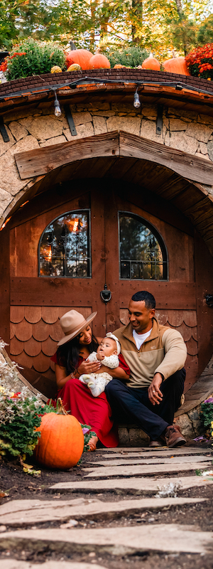 A family sits in front of a hobbit-style door, surrounded by pumpkins and autumn flowers. The setting is vibrant and cozy.