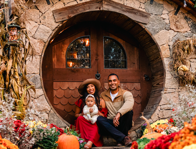 A family sits outside a hobbit-style house surrounded by pumpkins and colorful flowers.