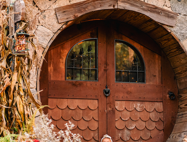 A small child sits in front of a round Hobbit-style door surrounded by autumn decorations, including pumpkins and colorful foliage.