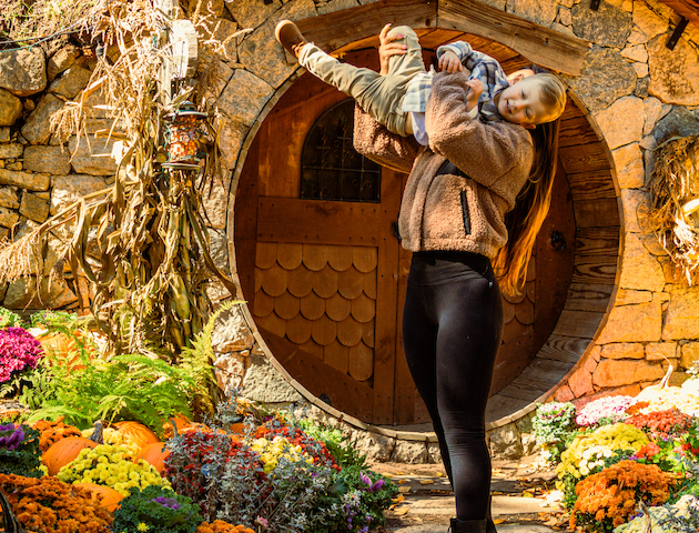 A person holds a child in front of a round, rustic door adorned with pumpkins and colorful flowers, creating a cozy, autumnal scene.