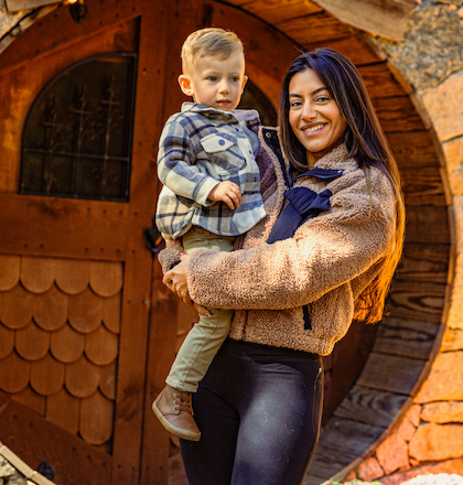 A person is holding a child in front of a round wooden door with a rustic design, surrounded by stone and wood elements.