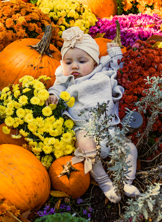 A baby dressed in a cozy outfit lies among vibrant flowers and pumpkins, enjoying an autumn scene.