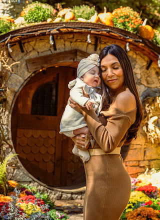 A woman holds a baby in front of a round wooden door, surrounded by colorful flowers and pumpkins.
