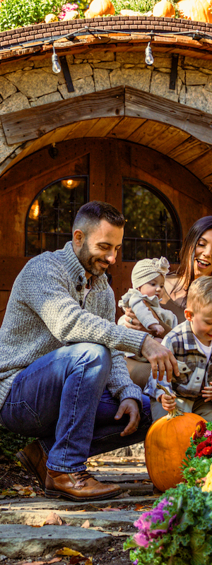 A family is gathered at a quaint, round door surrounded by lush flowers and pumpkins, enjoying a cheerful outdoor moment.