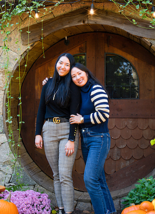 Two people stand smiling in front of a round wooden door, surrounded by pumpkins and foliage, in a cozy outdoor setting.