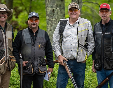 Four men in shooting vests and gear posing with shotguns in a scenic, wooded area at The Preserve Sporting Club, highlighting outdoor activities