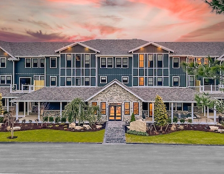 A large, three-story building with a stone entrance is surrounded by landscaped greenery against a vibrant sunset sky.