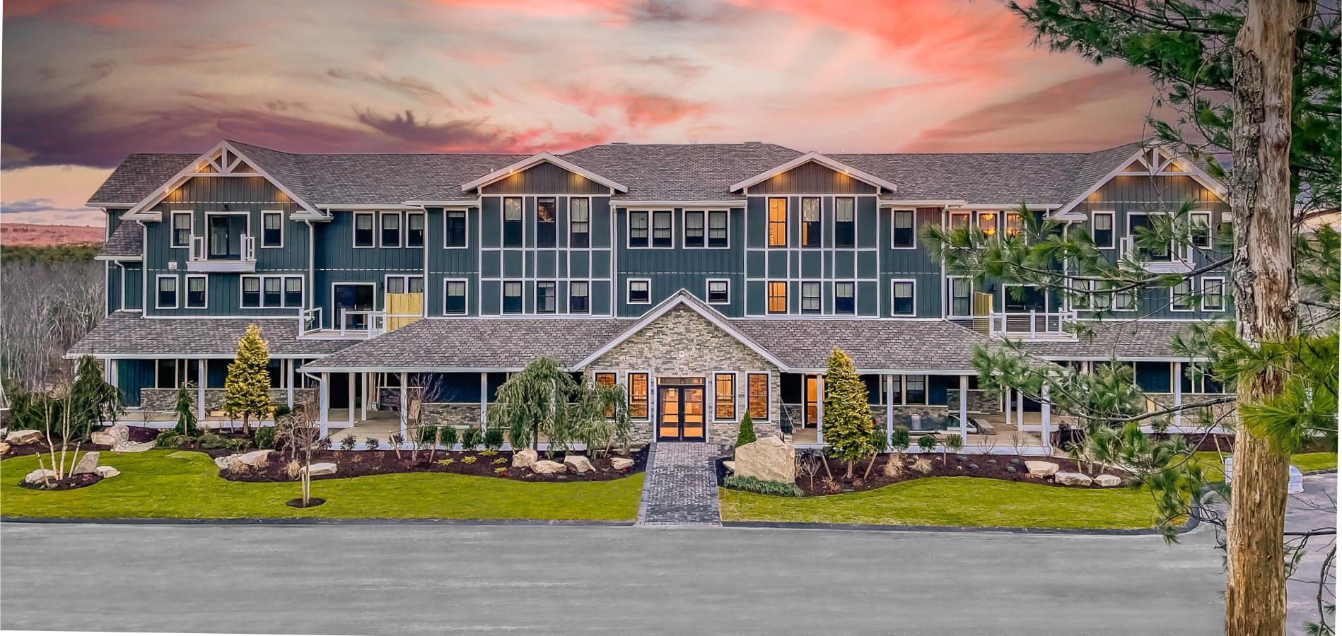 A large, three-story building with a stone entrance is surrounded by landscaped greenery against a vibrant sunset sky.