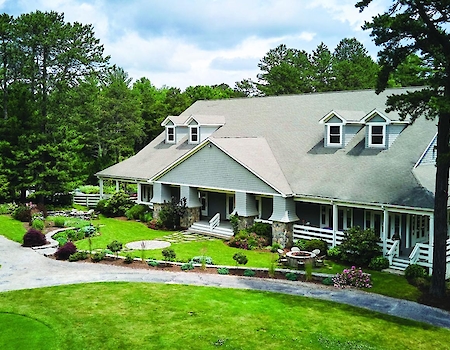 A large house with a wraparound porch is surrounded by well-maintained lawns and tall trees under a slightly cloudy sky. Elegant Venue at The Preserve Sporting Club