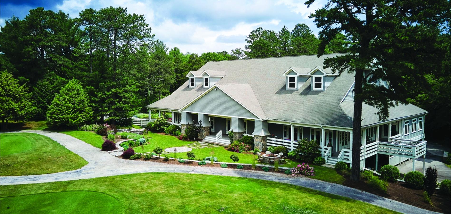 A large house with a wraparound porch is surrounded by well-maintained lawns and tall trees under a slightly cloudy sky. Elegant Venue at The Preserve Sporting Club