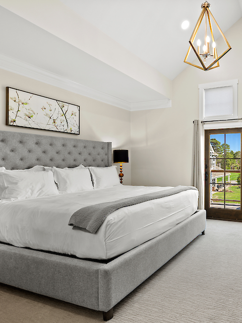 A spacious bedroom featuring a large bed, elegant armchair, chandelier, and glass doors leading to a scenic outdoor view.
