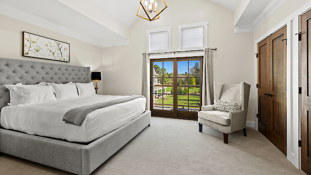 A spacious bedroom featuring a large bed, elegant armchair, chandelier, and glass doors leading to a scenic outdoor view.