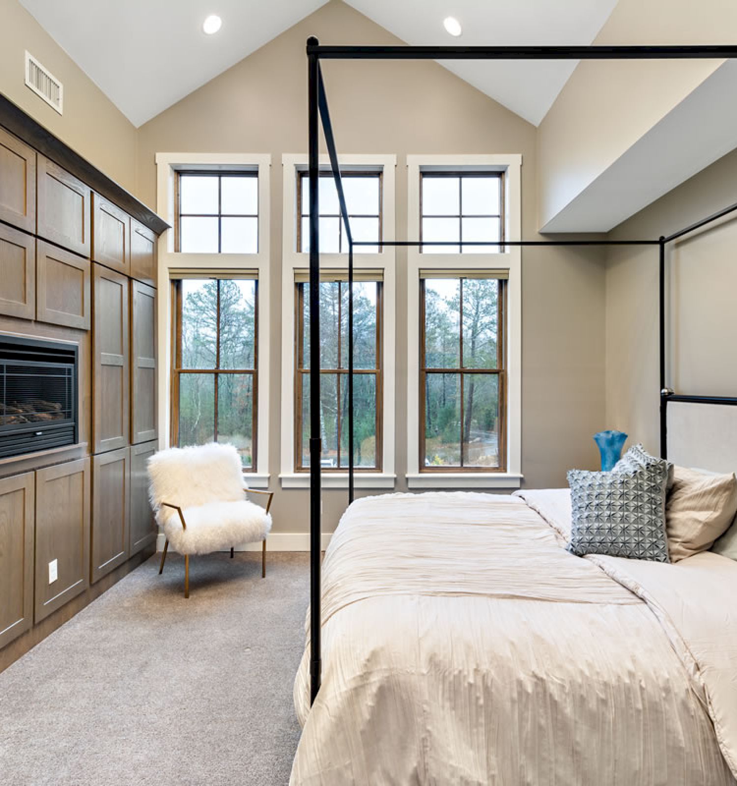 A modern bedroom with a canopy bed, large windows, built-in wooden cabinets, a fireplace, and a white chair near the windows, neutral d&eacute;cor.
