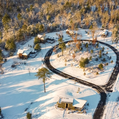 An aerial view of a snowy landscape with scattered cabins and surrounding trees, featuring a curved road leading through the area.
