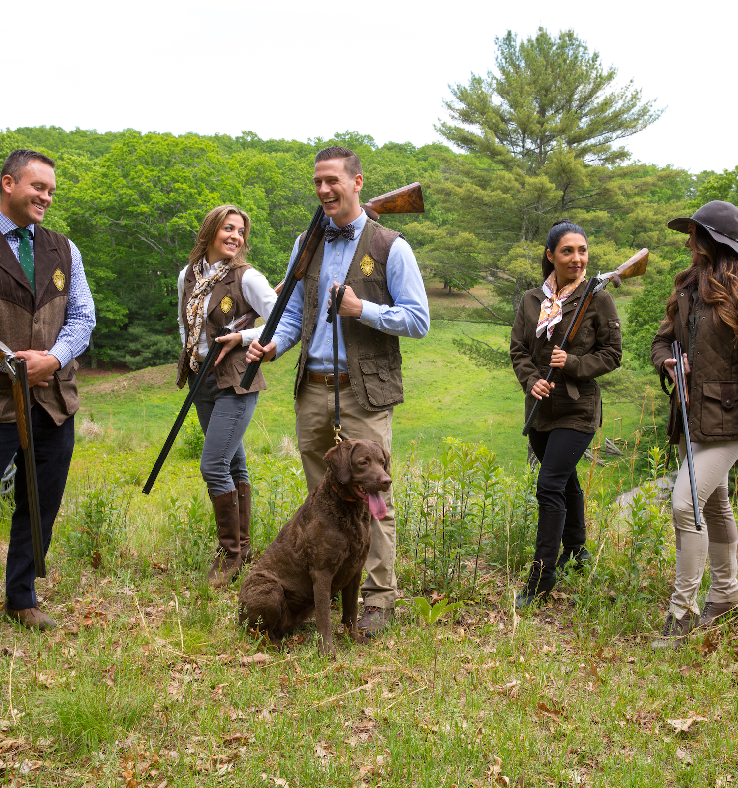 A group of people with rifles and a dog stand outdoors in a grassy area, dressed in hunting attire, with trees and a vehicle in the background.