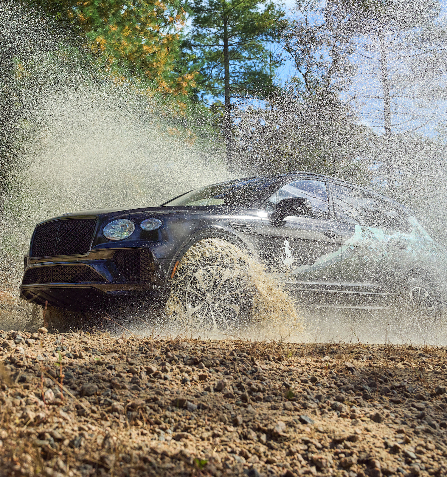 A car is driving through a muddy, rocky terrain, splashing water, with trees in the background under a clear sky.