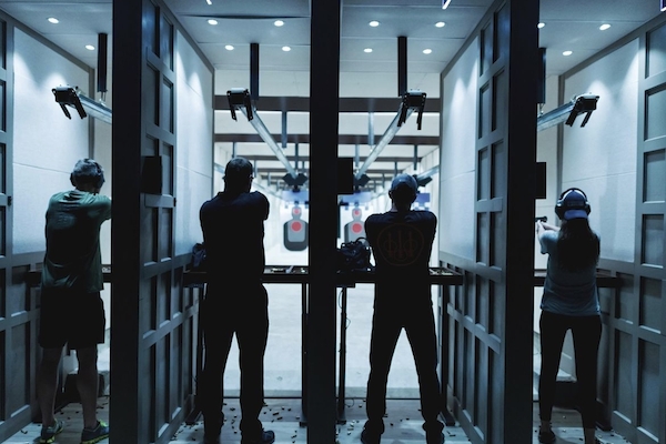 The image shows four people in individual shooting stalls at an indoor firing range, aiming at targets.