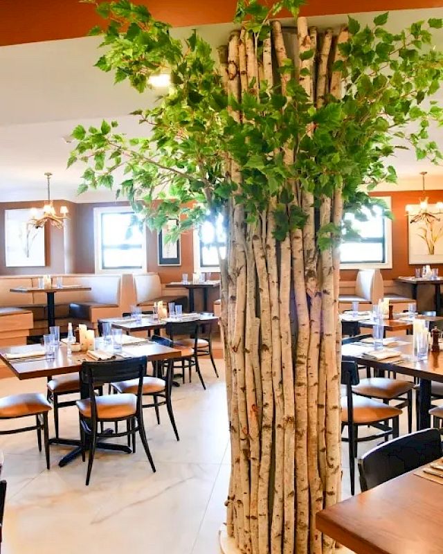 A restaurant interior with wooden tables and chairs, featuring a decorative tree installation in the center surrounded by cozy dining areas.