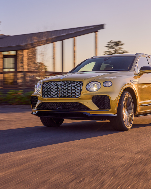 A luxury SUV is driving on a sunlit road, with a modern house and trees in the background.