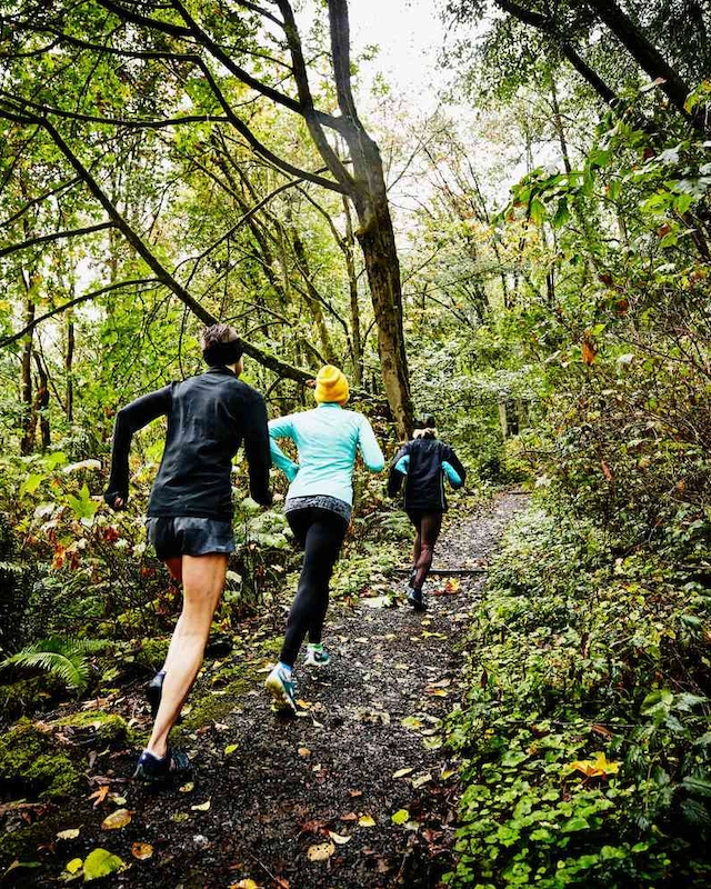 Three people are running on a forest trail surrounded by lush trees and greenery. The path is slightly muddy, indicating recent rain.