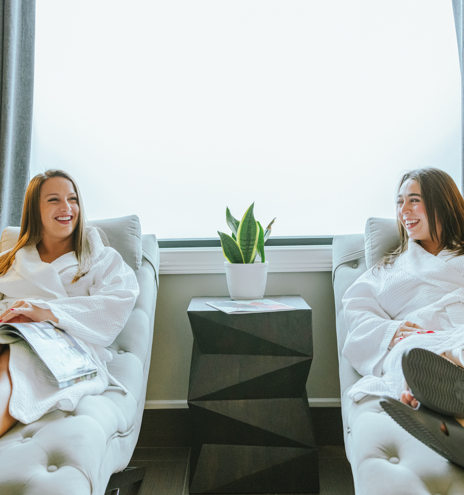 Two people in bathrobes relax on lounge chairs with drinks, a plant between them, against a bright window.