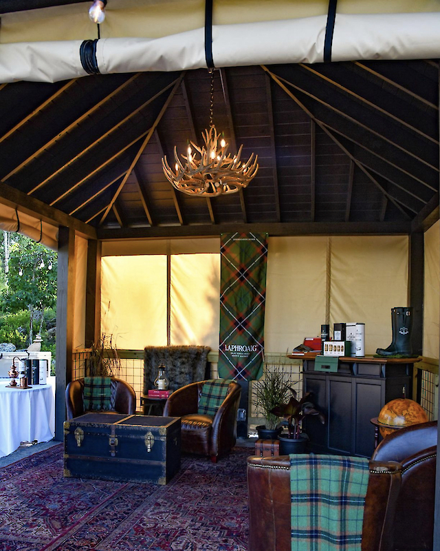 A cozy tent with a chandelier, armchairs, trunk table, and plaid decor is depicted, creating a rustic, inviting atmosphere inside.