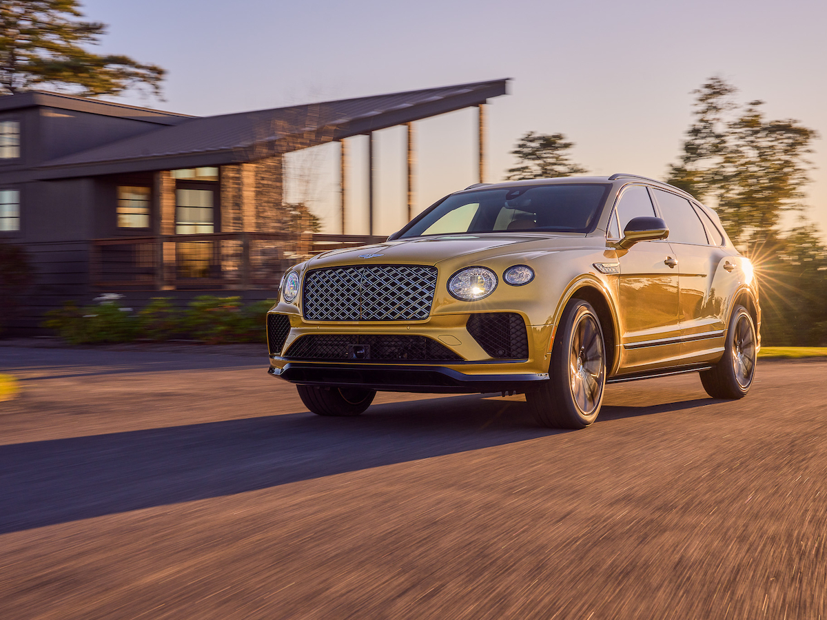 A gold luxury SUV is driving on a road near a modern house with a sunset backdrop.
