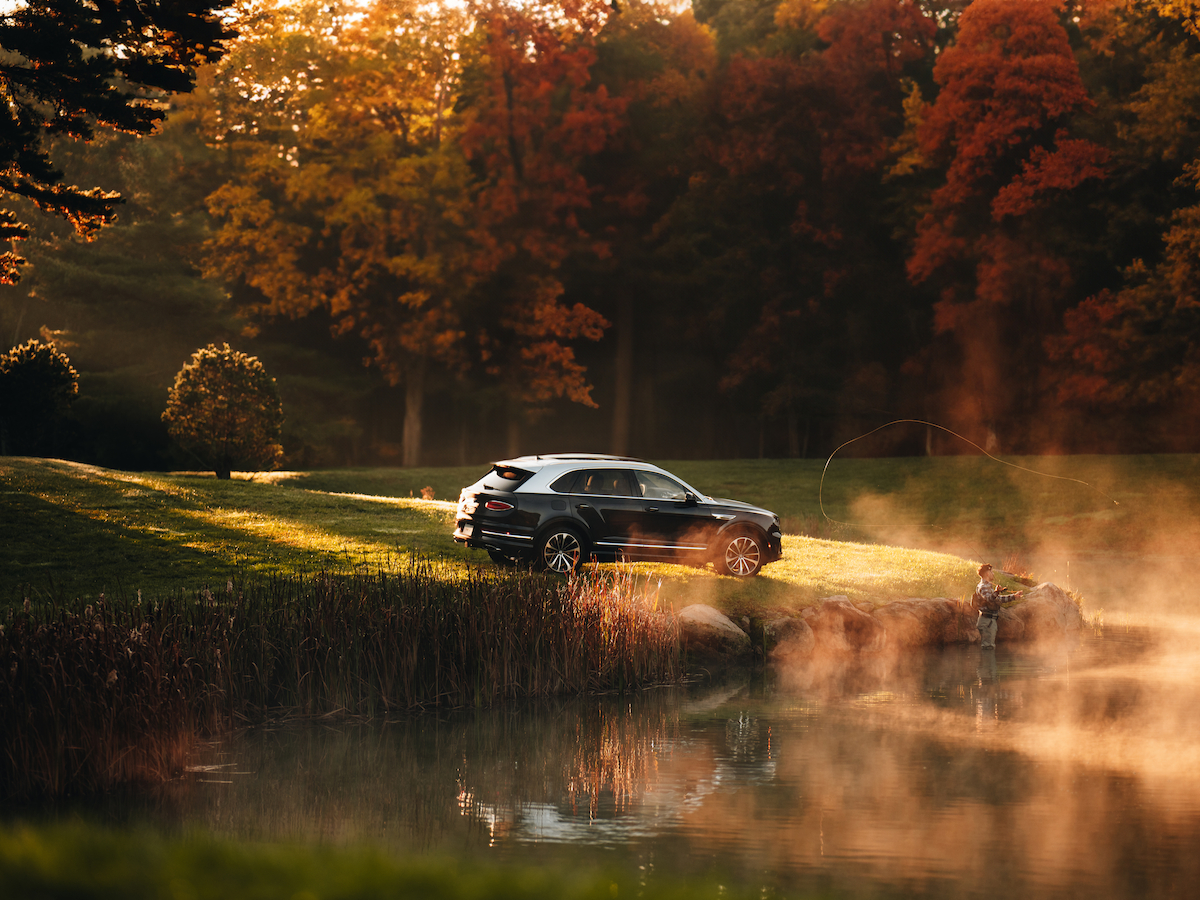 A car is parked beside a misty pond surrounded by autumn trees, with sunlight filtering through the foliage, creating a serene scene.