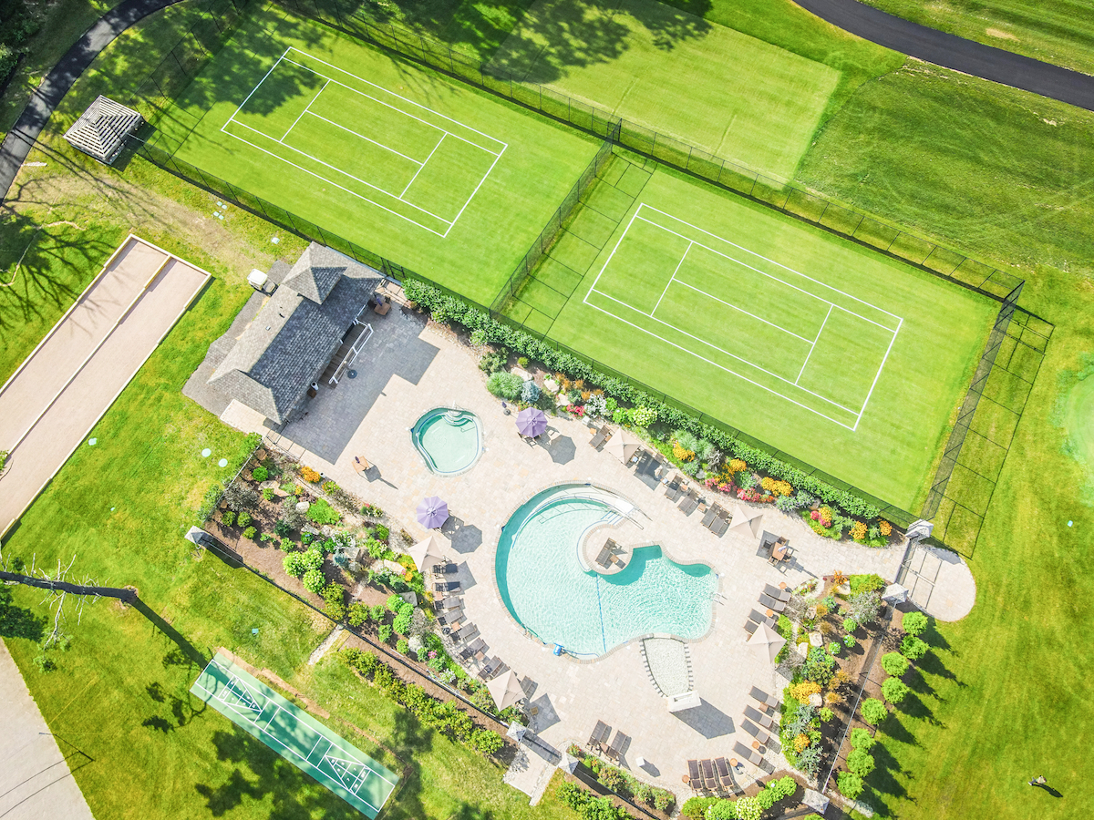 This image shows two tennis courts and a landscaped area with a large pool, lounge chairs, and surrounding greenery from an aerial view.