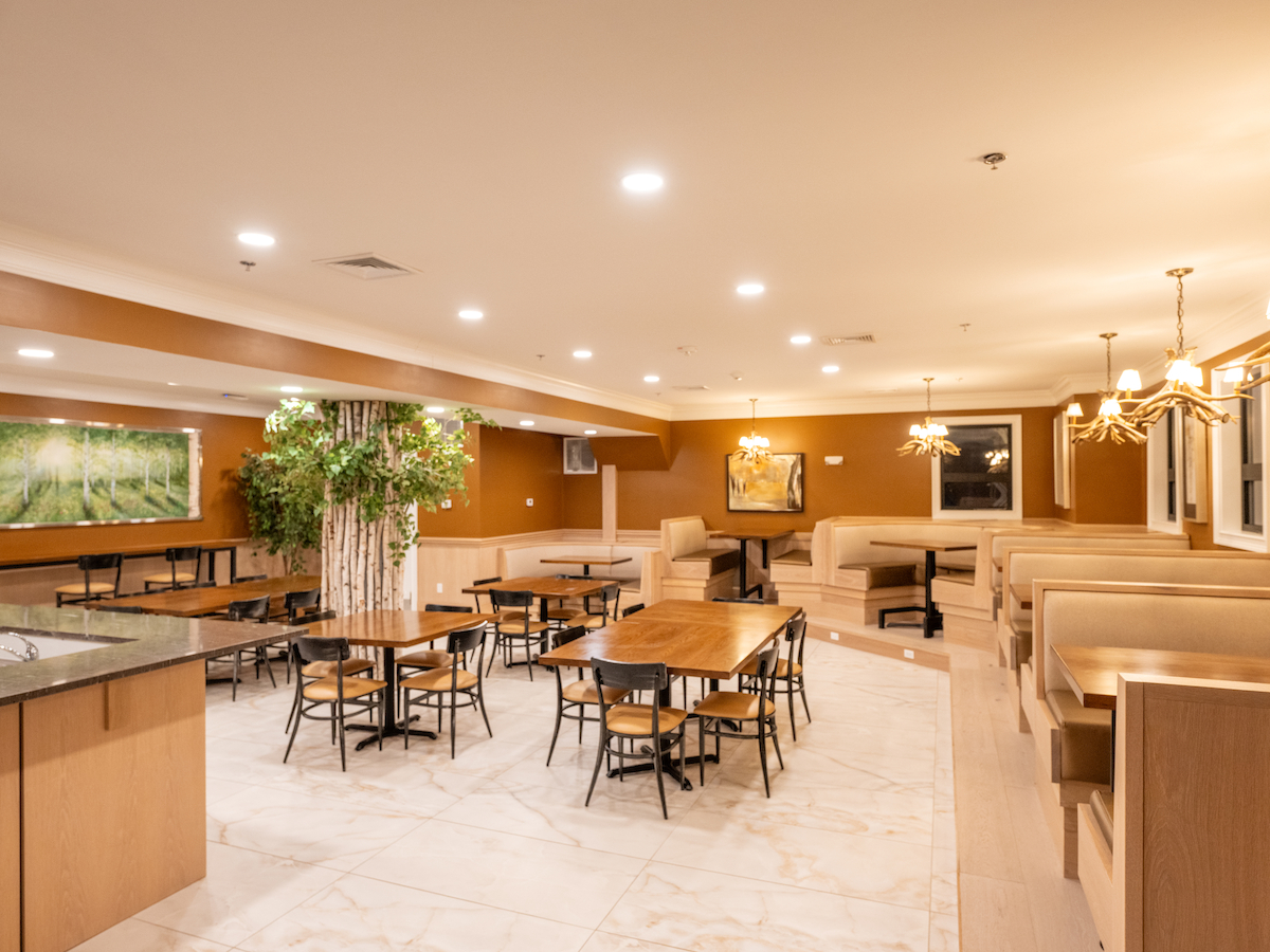 A spacious and well-lit restaurant interior with wooden tables, chairs, and booths, featuring decorative plants and framed artwork.