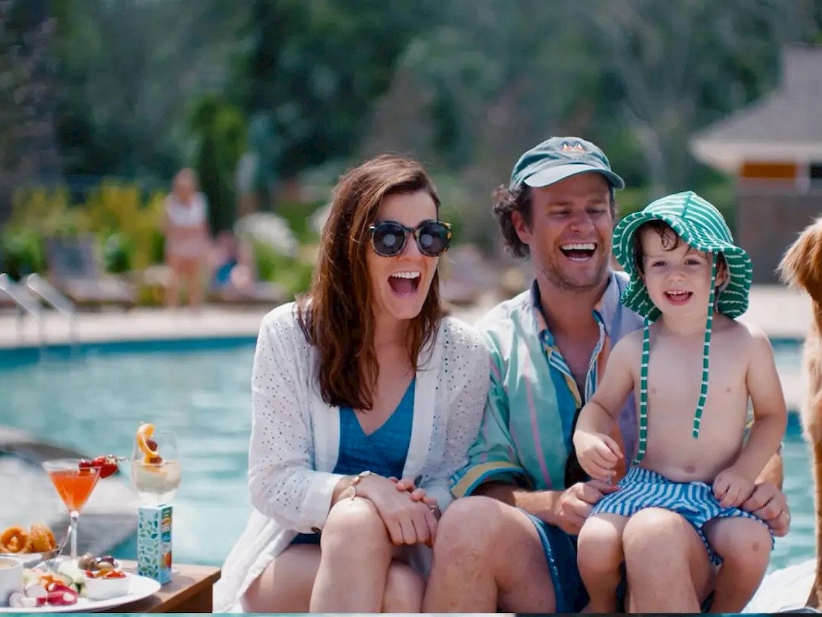 A group by a pool: two adults, a child in a hat, and a dog. They're smiling with snacks and drinks on a nearby table.
