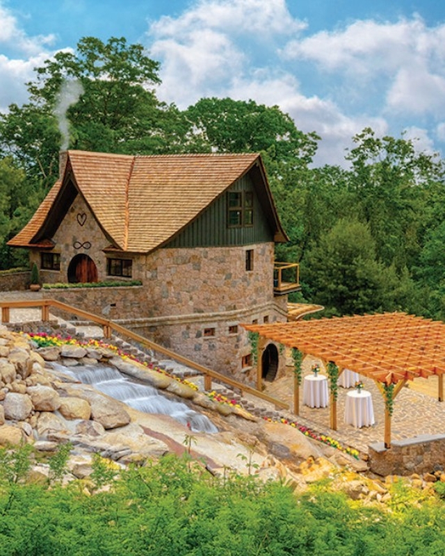 A charming stone cottage with a water feature and patio area is surrounded by lush greenery and a blue sky dotted with clouds.