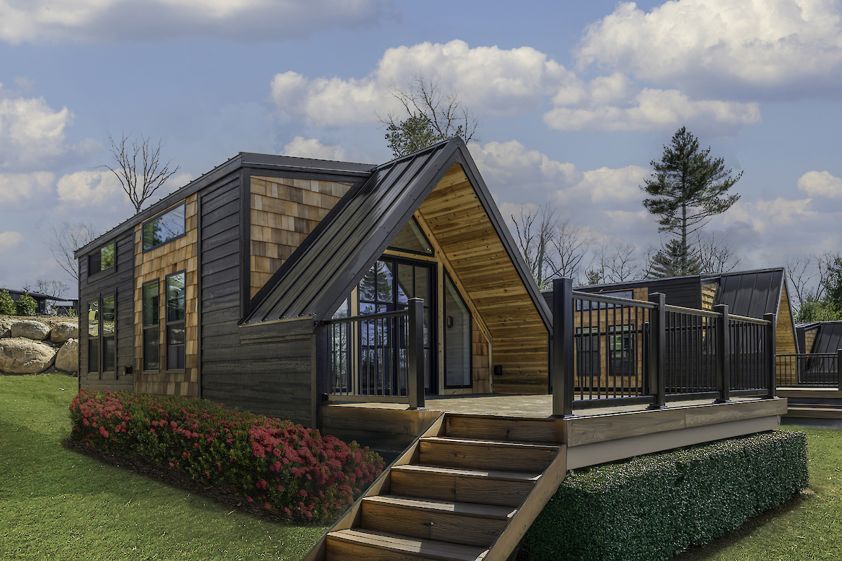 A small modern cabin with wooden finishes sits on a porch, surrounded by grass and flowers, under a cloudy sky.
