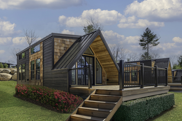 A modern tiny house with wooden siding, patio, steps, and a grassy yard, under a partly cloudy sky.