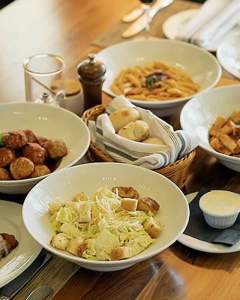 Assorted dishes including pasta, meatballs, salad with croutons, bread rolls, and a small bowl of sauce on a wooden table.