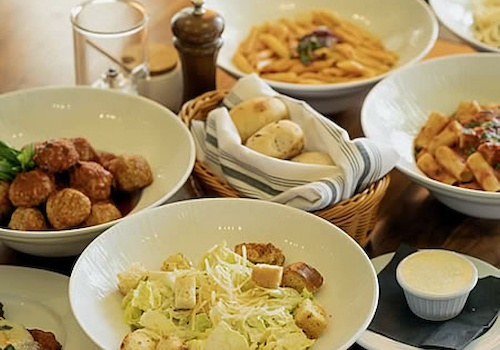 Assorted dishes including pasta, meatballs, salad with croutons, bread rolls, and a small bowl of sauce on a wooden table.